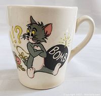 Front view showing Tom sitting near a bomb with 'BOMB' text and Jerry's surprised face with exclamation marks on a white ceramic mug.