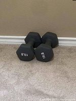 Image showing two black hexagonal 15 lbs dumbbells resting on carpeted floor against a beige wall.