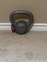 Photo showing a dark gray 15 lbs cast iron kettlebell with a wide handle, standing upright against a wall on carpeted floor.