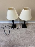 Two oiled bronze table lamps with cream fabric lampshades, cords attached, placed against a beige wall.