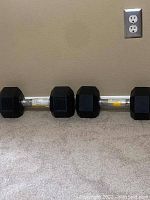 Photo showing two 25 pound hex dumbbells placed on carpeted floor against a beige wall with an electrical outlet visible.