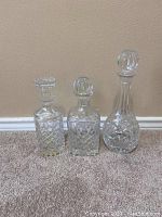 Photo showing three clear glass decanters with stoppers on carpeted floor against beige wall. Decanters include two square shaped with cut patterns and one taller rounded one.