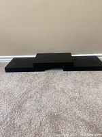 Three black floating shelves arranged on a carpeted floor showing a stepped design with a raised center shelf and two flat side shelves.
