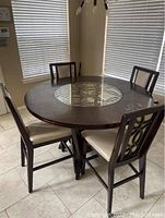 Four counter height dining chairs with wooden dark finish frames and light cushioned seats arranged around a round wooden dining table with glass inset.