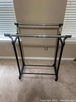 Photo of an adjustable metal clothing rack with two horizontal chrome bars and caster wheels on a carpeted floor.