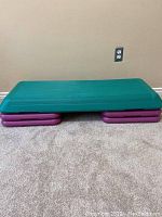 Green aerobic step platform resting on four purple risers on floor in front of beige wall.