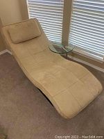 Tan suede-like chaise lounge with an attached pillow and round glass side table with metal pedestal base, placed near window with blinds.