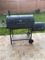Full view of the black charcoal grill with attached smoker box, side shelf, thermometer on lid, and wheels, placed on tiled outdoor floor.
