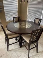 Round counter height dining table shown with four chairs for scale only, chairs not included in the sale. Table has dark wood finish and a glass center insert with decorative design.