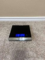 Digital food scale showing blue backlit LCD screen with weight reading on carpeted floor.