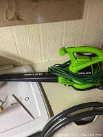Green Portland electric leaf blower with a long power cord wrapped around the handle and black nozzle attachment placed on a white surface against a textured wall.
