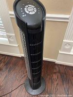 Black tower fan standing on hardwood floor near window with blinds. Fan has vertical slats and control buttons on top.