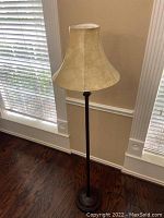 Floor lamp positioned between two windows with wooden flooring as background. Lamp has a beige fabric bell-shaped lampshade and dark metal stand with round base.