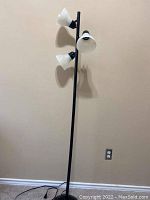 Black metal floor lamp with three white frosted glass lampshades attached vertically along the pole against a beige wall.