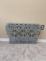 Gray ornate wall decor panel with decorative scroll and floral pattern leaning against beige wall next to an electrical outlet.