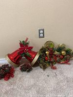 Photo showing two large red jingle bells with gold rims and a festive garland with pinecones, berries, and ribbons on the floor against a beige wall.