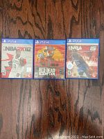 Three PS4 game cases, NBA 2K18, Red Dead Redemption II, and NBA 2K15, laid flat on dark wood surface