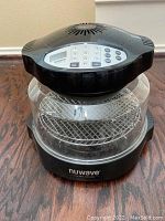 Black Nuwave Oven with digital control panel, two metal wire racks inside, and glass cooking bowl shown on wood floor.