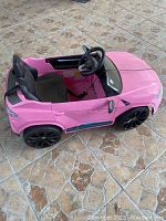 Pink kids electric toy car resembling a Porsche, single-seat, black seat and steering wheel, black rims, on tiled floor.