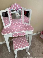 Pink kids vanity table with white polka dots on the pink surface, tri-fold mirrors, and matching stool in front.