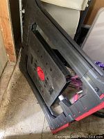 Folded portable work bench with black and red plastic frame, purple clamps attached underneath, stored on floor next to chair.