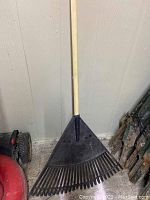 Photo of a black plastic leaf rake head on a wooden handle leaning against a light wall with some other garden items nearby.
