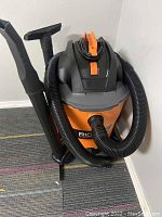 Orange and black Rigid wet/dry vacuum cleaner standing on carpeted floor in a corner. Hoses and attachments are connected and arranged on the unit.
