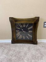 Square wall clock with dark wooden frame and Roman numeral face on a beige wall.