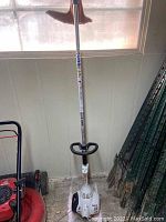 Photo of the full Stihl gas powered weed eater standing vertically inside a room next to a red lawn mower and some green garden stakes.