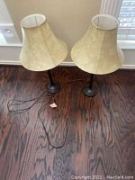 Two brown table lamps with beige bell-shaped lampshades and visible cords on a wooden floor against a wall.