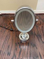 Chrome oval vanity mirror with LED light ring on a round base with adjustable tilt, plugged in with power cord visible.