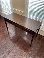Brown wooden rectangular desk with visible scratches on the surface, placed on dark hardwood floor near window blinds.
