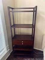 Brown wooden shelf with three open shelves and one drawer in middle with two knobs.