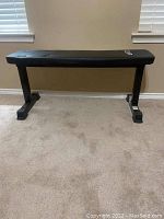 Photo of a black flat workout bench against a beige wall and carpeted floor. The bench has a black padded seat with a 'GP' logo and metal legs with rubber feet.