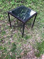 Accent table shown outdoors on grass with black metal frame and square mirrored glass top reflecting sky and tree branches.