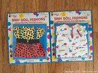 Photo showing two Baby Doll Fashions outfits in original packaging, one with yellow and red polka dots, the other white with colorful shapes.