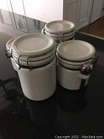 Four white ceramic canisters with metal clamp lids arranged together on a surface, seen from above at an angle.