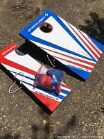 Two Sportcraft cornhole boards with red, white, and blue triangular stripes, foldable legs, and eight red and blue bean bags in a clear bag placed on one board