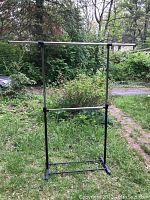 Photo showing the full black metal double hanger clothing rack standing outside on grass with trees in background.