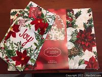 Overview of 2 folded kitchen towels on 4 placemats in original packaging, showing holiday red, green and white poinsettia and holly designs