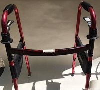 Red folding walker with black plastic handles and adjustable height, showing front view in sunlight on concrete.