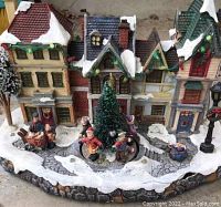 Front view of detailed European village Christmas scene featuring snow-covered buildings, a green twirling Christmas tree, and children figurines in winter attire.