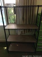 Black 4-tier metal wire shelving unit with wood or wood-like shelves against a window and curtains background.