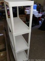 Photo showing the white plastic shelving unit featuring four tiers and cylindrical vertical supports, placed in a garage or storage setting.