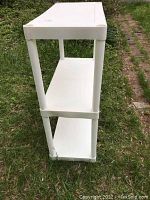 Side angled view showing three white plastic shelves with cylindrical supports, outdoors on grass.