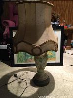 Full view of the vintage lamp showing the porcelain floral base and fabric lampshade on a carpeted floor with some background items.