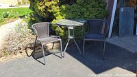 Bistro set with two brown wicker chairs and round glass topper table on metal frame outside near bushes.