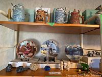 Full lot view showing three decorative plates on stand, four beer steins on upper shelf, and small figurines and objects on lower shelf.