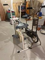 Two stationary exercise bikes situated in a basement area; one beige with an electronic display and fan, the other a black vintage model.