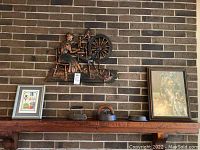 View of a dark wooden mantle shelf with metal wall art of a woman spinning thread mounted on brick wall and an array of iron cast pieces and framed art on shelf